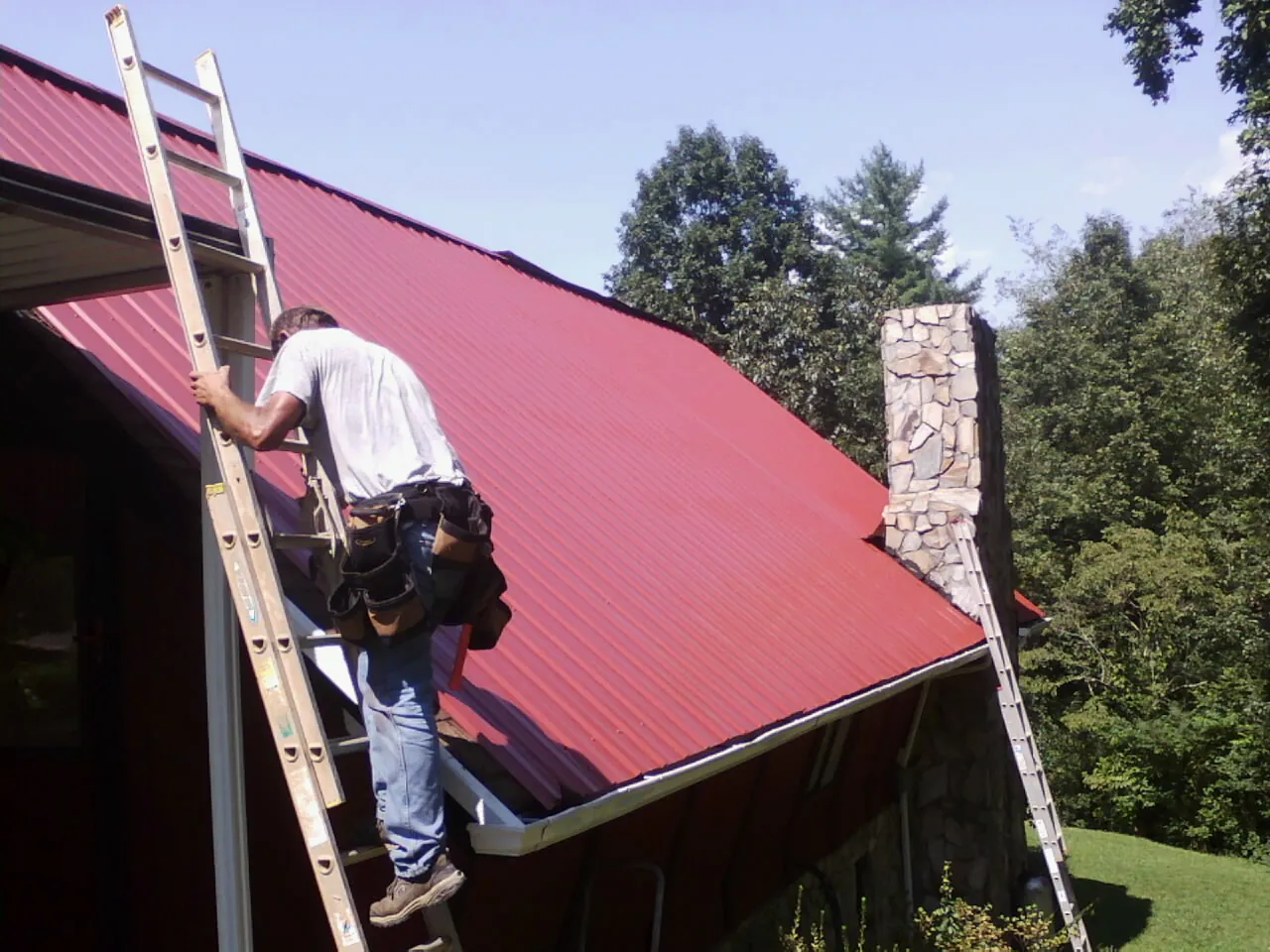 Roofer on a ladder beside a red metal roof and stone chimney — Soffit & Fascia Repair by Roofing Brothers in Basking Ridge